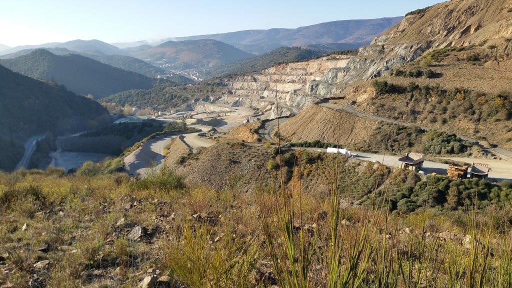 Vue panoramique d'un paysage de montagne avec une carrière à ciel ouvert et une route sinueuse, entourée de végétation.