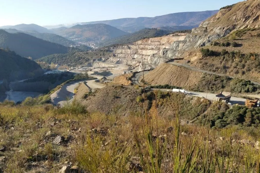 Vue panoramique d'un paysage de montagne avec une carrière à ciel ouvert et une route sinueuse, entourée de végétation.