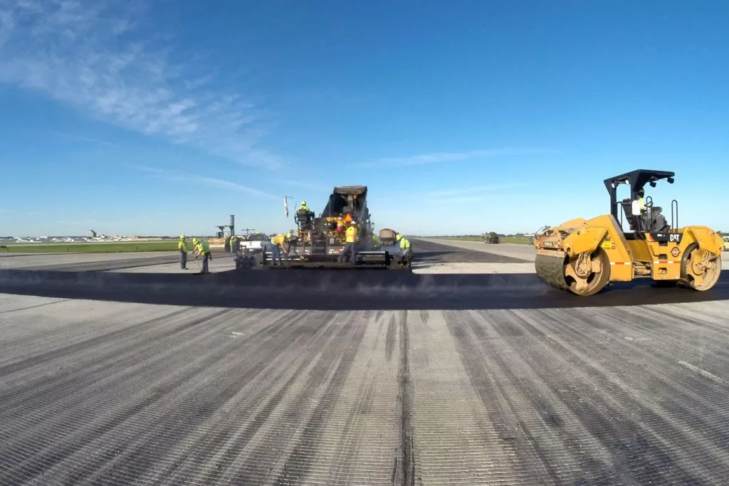 Travaux de construction sur une piste d'aéroport avec un rouleau compresseur et une équipe utilisant une machine, sous un ciel bleu.