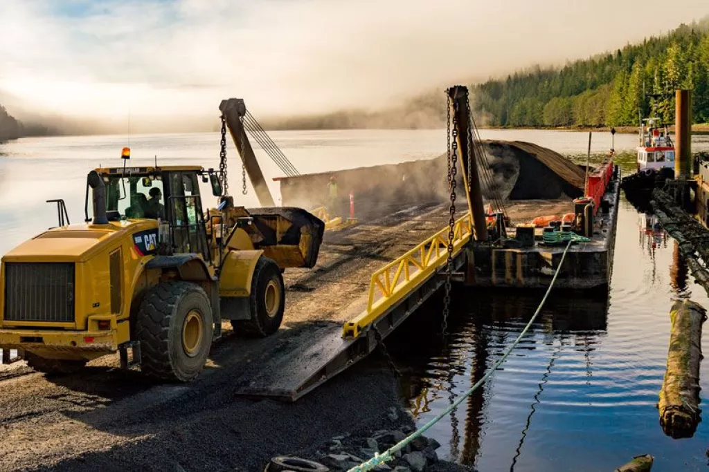 Chantier de construction au bord de l'eau avec un bulldozer déplaçant de la terre sur une péniche, entouré de forêt et de montagnes embrumées.