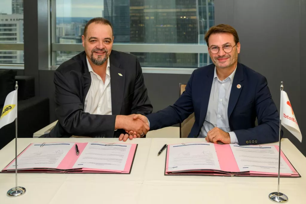 Deux représentants de Colas et de TotalEnergies se serrent la main après la signature d’un accord de partenariat, assis à une table où figurent les documents signés et les drapeaux des deux entreprises.