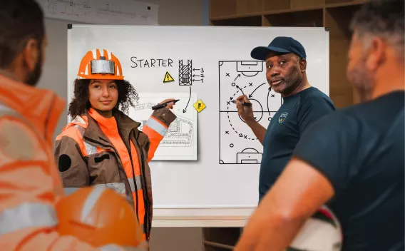Animatrice Colas en tenue de chantier et coach sportif expliquent sur un tableau la stratégie de préparation sécurité pendant la Safety Week 2026.