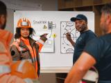 Animatrice Colas en tenue de chantier et coach sportif expliquent sur un tableau la stratégie de préparation sécurité pendant la Safety Week 2026.