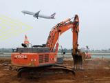 Pelle mécanique en activité sur un chantier aéroportuaire, avec un avion au décollage en arrière-plan
