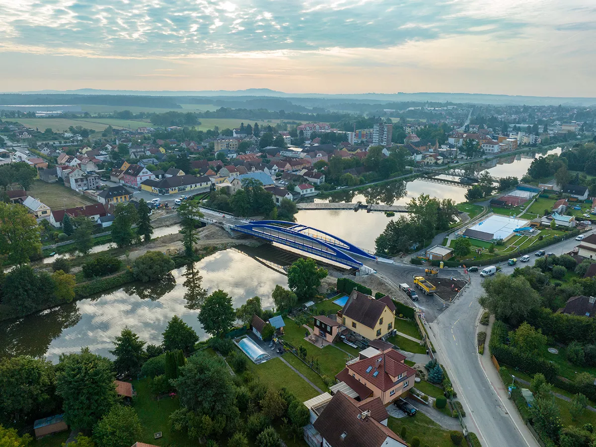 Aerial view of a newly installed blue bridge over a river, in the heart of a residential town surrounded by greenery and roads under construction.