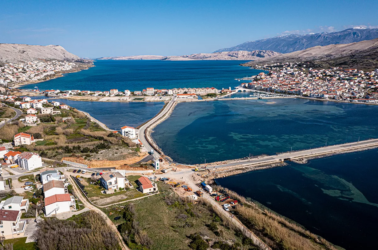 Aerial view of an island connected by roads surrounded by water, with a coastal village and mountains in the background.