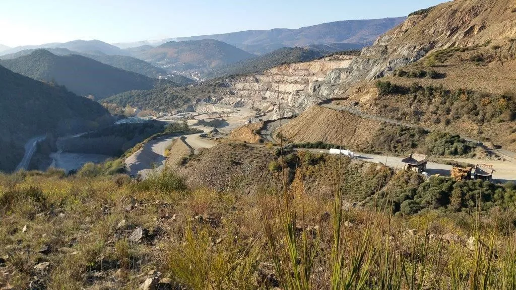 Panoramic view of a mountain landscape with an open quarry and a winding road, surrounded by vegetation.
