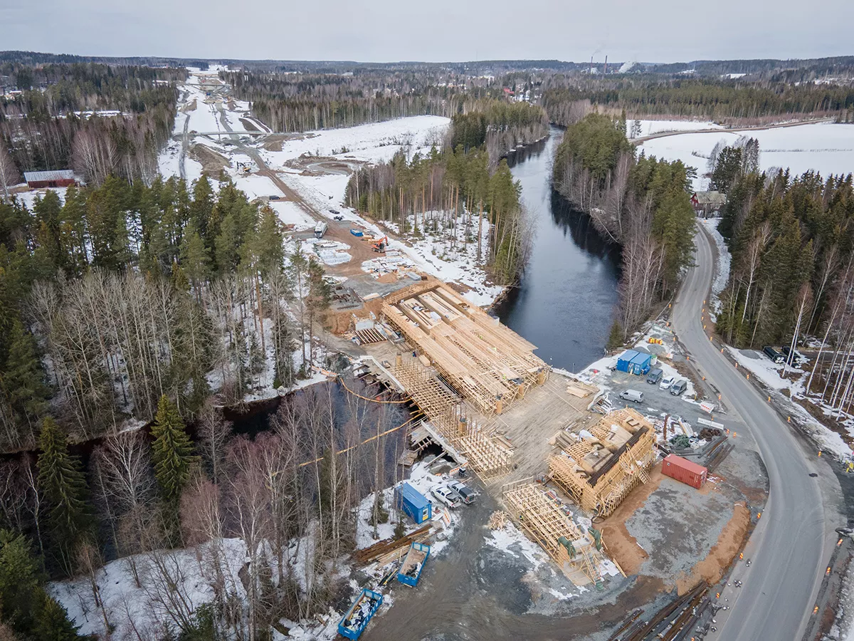 Construction site of a wooden bridge over a river surrounded by a forest covered with snow