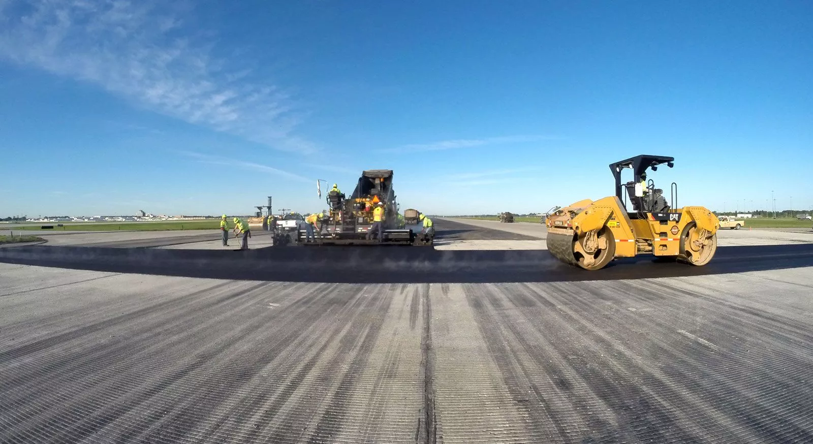 Construction work on an airport runway with a steamroller and a team using a machine, under a blue sky.