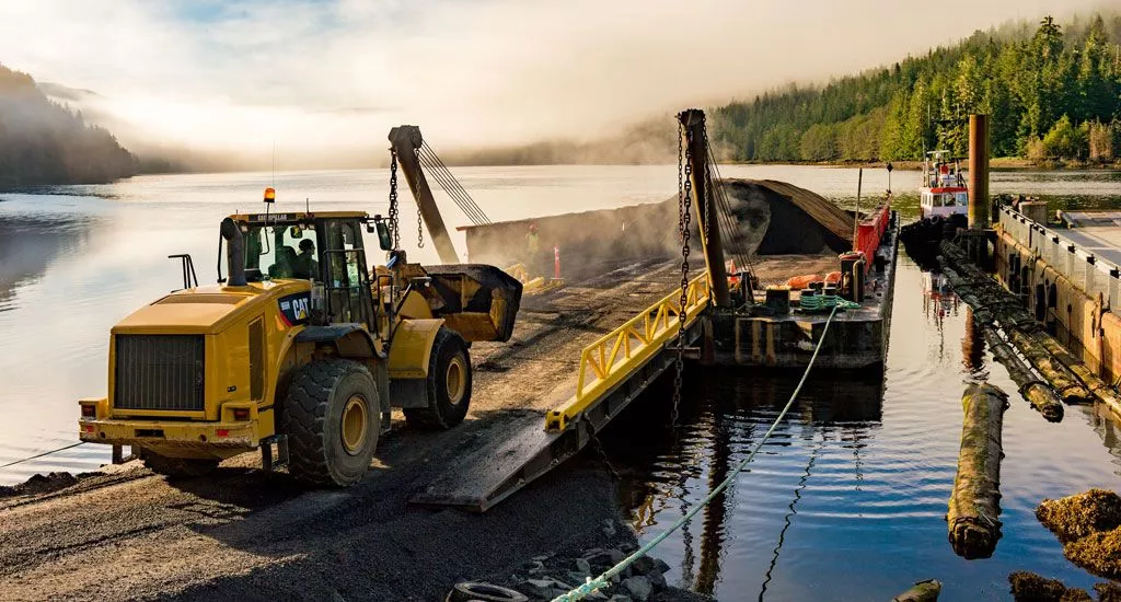 Construction site at the edge of the water with a bulldozer moving land on a barge, surrounded by forest and misty mountains.