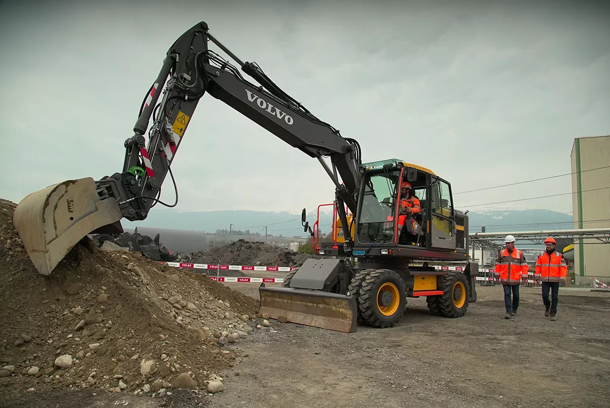 Volvo excavator in the construction site, operator on board, two workers in orange uniform are talking next to each other