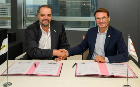 Deux représentants de Colas et de TotalEnergies se serrent la main après la signature d’un accord de partenariat, assis à une table où figurent les documents signés et les drapeaux des deux entreprises.