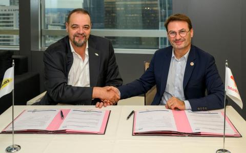Deux représentants de Colas et de TotalEnergies se serrent la main après la signature d’un accord de partenariat, assis à une table où figurent les documents signés et les drapeaux des deux entreprises.